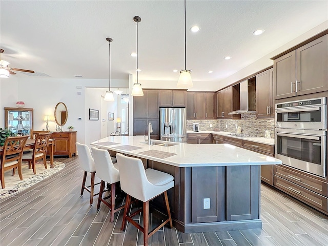 kitchen featuring appliances with stainless steel finishes, a kitchen island with sink, hanging light fixtures, backsplash, and wall chimney exhaust hood