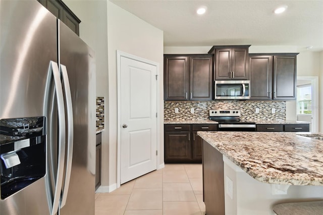 kitchen with appliances with stainless steel finishes, backsplash, light tile patterned floors, dark brown cabinetry, and light stone countertops