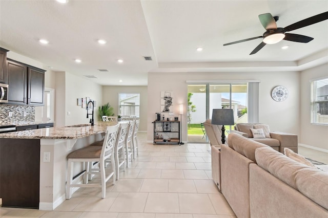 kitchen with a breakfast bar area, backsplash, a kitchen island with sink, ceiling fan, and light stone countertops