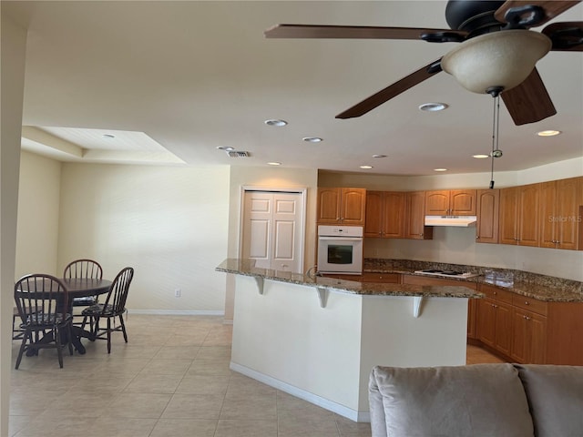 kitchen featuring light tile patterned flooring, a breakfast bar area, dark stone countertops, white appliances, and ceiling fan