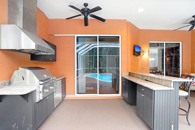 kitchen with a breakfast bar, wall chimney exhaust hood, light stone counters, ceiling fan, and refrigerator