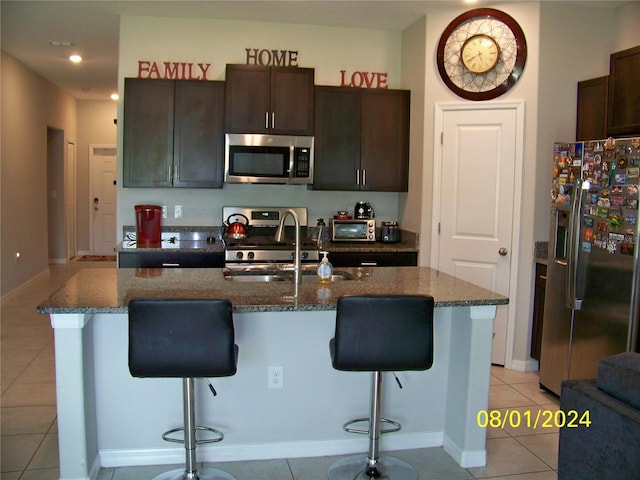 kitchen featuring dark brown cabinetry, stainless steel appliances, a center island with sink, a breakfast bar area, and light tile patterned flooring