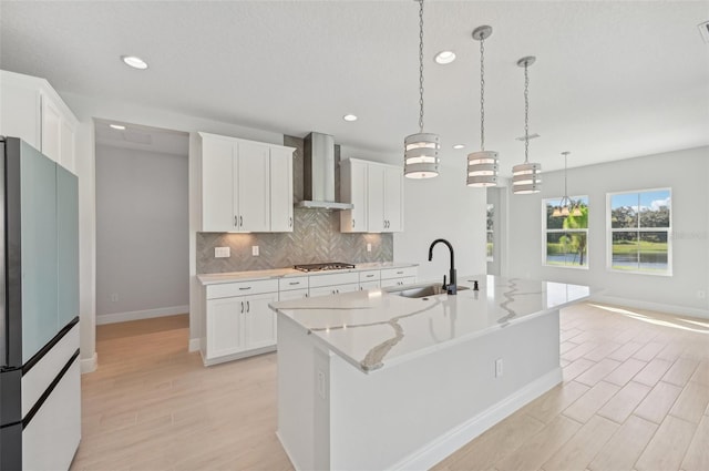 kitchen with wall chimney range hood, sink, a kitchen island with sink, white cabinetry, and fridge