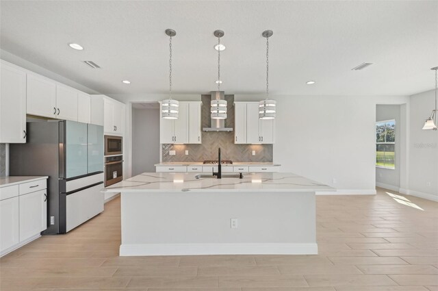 kitchen featuring appliances with stainless steel finishes, white cabinetry, an island with sink, light stone counters, and wall chimney exhaust hood