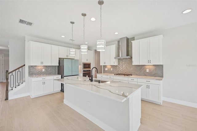 kitchen featuring appliances with stainless steel finishes, white cabinetry, sink, a kitchen island with sink, and wall chimney range hood