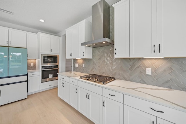 kitchen featuring wall chimney exhaust hood, white cabinetry, light wood-type flooring, appliances with stainless steel finishes, and light stone countertops