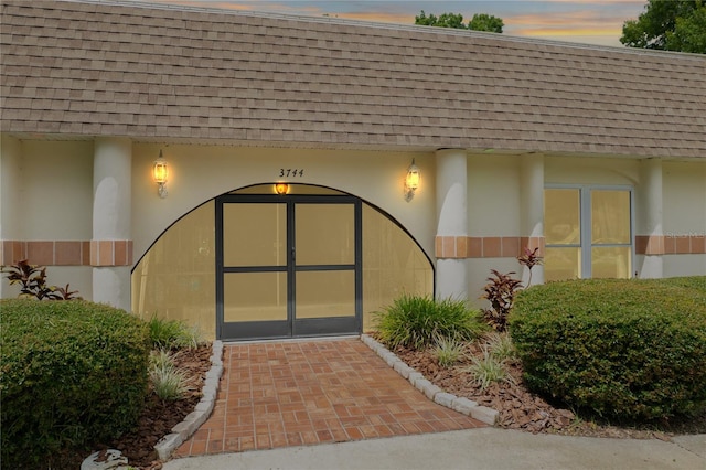 exterior entry at dusk with stucco siding, mansard roof, and roof with shingles