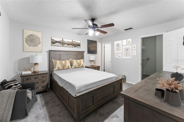 bedroom with a spacious closet, a textured ceiling, a closet, ceiling fan, and dark carpet