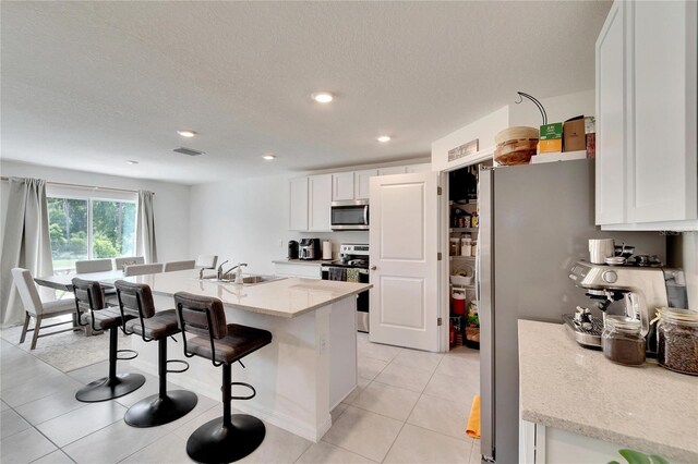 kitchen featuring appliances with stainless steel finishes, white cabinets, light tile patterned flooring, a center island with sink, and sink