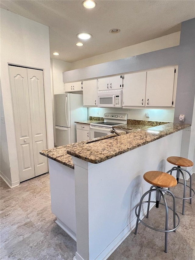 kitchen featuring white cabinetry, dark stone countertops, a kitchen breakfast bar, kitchen peninsula, and white appliances