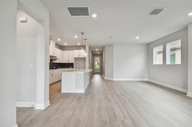 kitchen with decorative light fixtures, white cabinetry, backsplash, a center island with sink, and light hardwood / wood-style flooring