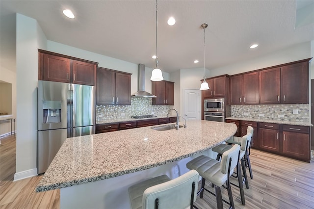 kitchen with wall chimney exhaust hood, sink, hanging light fixtures, appliances with stainless steel finishes, and a kitchen island with sink