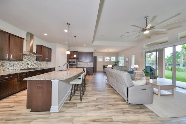 kitchen with wall chimney range hood, sink, a center island with sink, decorative light fixtures, and stainless steel gas stovetop