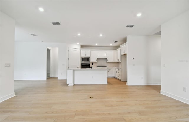 kitchen with an island with sink, light hardwood / wood-style floors, white cabinets, custom range hood, and appliances with stainless steel finishes