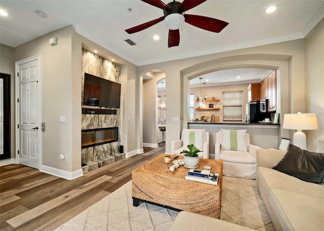 living room featuring ceiling fan, light hardwood / wood-style flooring, a stone fireplace, and ornamental molding