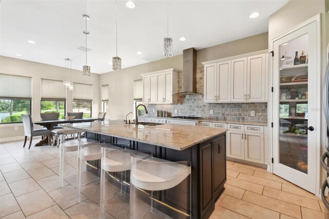 kitchen with an island with sink, backsplash, sink, wall chimney range hood, and decorative light fixtures