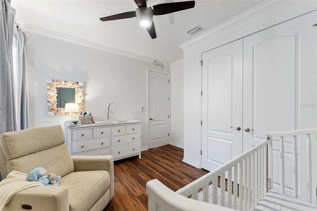 bedroom with ceiling fan, dark wood-type flooring, crown molding, a crib, and a closet