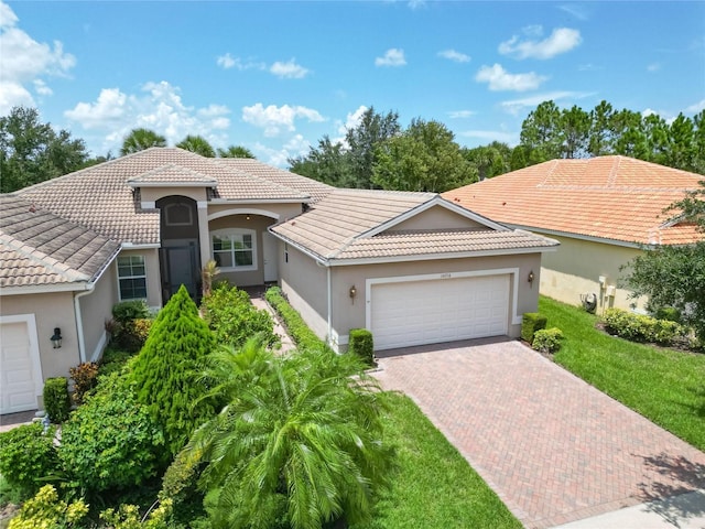 mediterranean / spanish-style house with an attached garage, a tiled roof, decorative driveway, stucco siding, and a front lawn