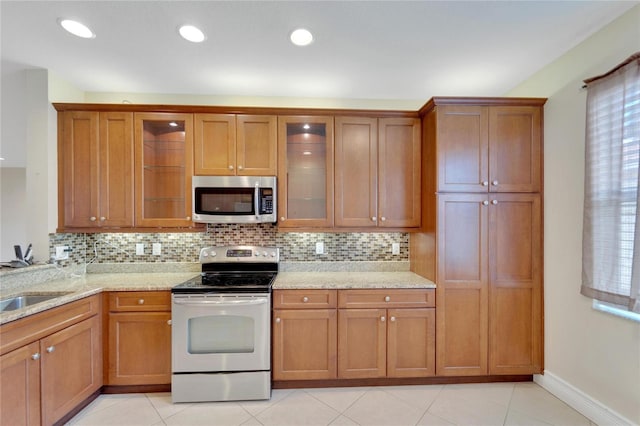 kitchen featuring brown cabinetry, glass insert cabinets, stainless steel appliances, and light stone counters