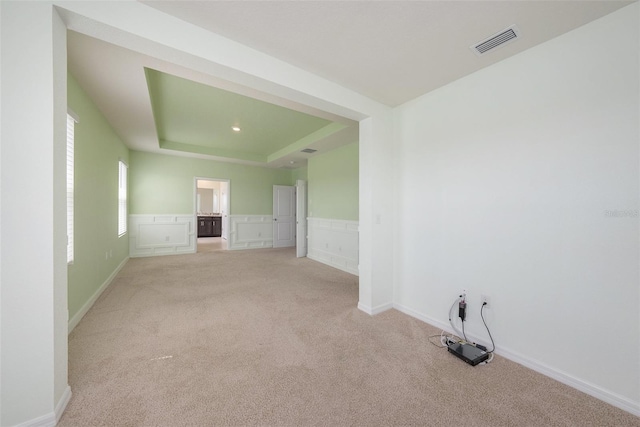 empty room with light carpet, visible vents, a raised ceiling, wainscoting, and a decorative wall