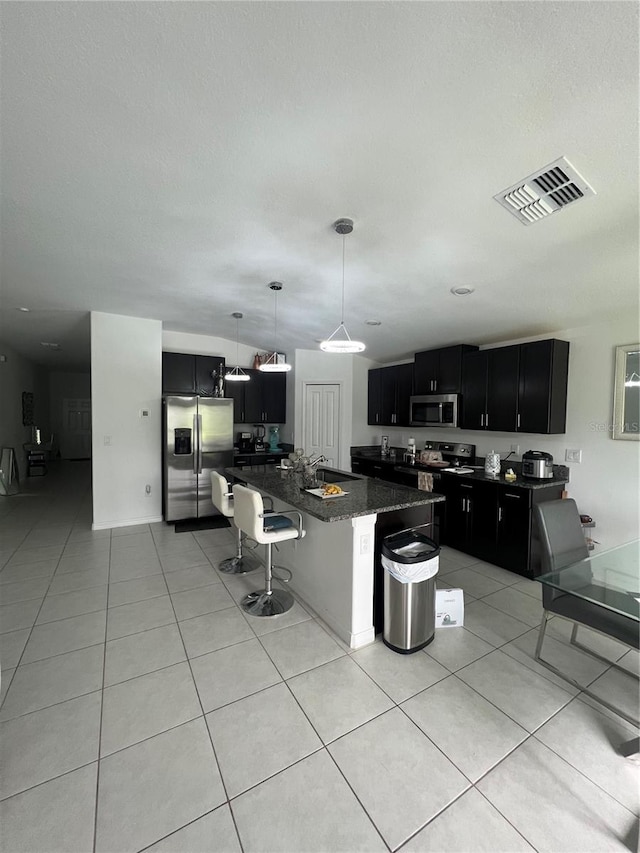 kitchen with appliances with stainless steel finishes, a kitchen breakfast bar, decorative light fixtures, dark stone countertops, and a center island with sink