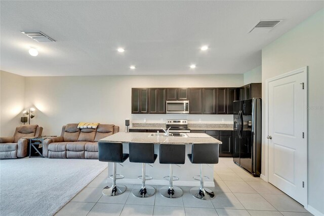 kitchen with a kitchen breakfast bar, dark brown cabinets, light tile patterned floors, stainless steel appliances, and a center island with sink