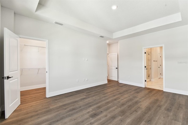 unfurnished bedroom featuring baseboards, visible vents, a tray ceiling, dark wood-type flooring, and a walk in closet