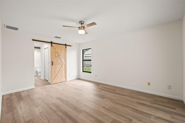 unfurnished bedroom with light hardwood / wood-style flooring, a barn door, ceiling fan, and ensuite bathroom