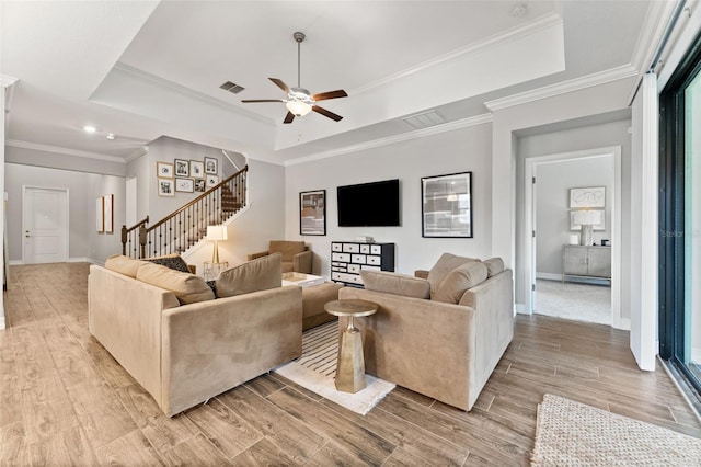 living room with light wood-type flooring, ornamental molding, ceiling fan, and a raised ceiling