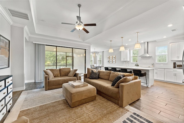 living room with ceiling fan, a raised ceiling, ornamental molding, light wood-type flooring, and sink