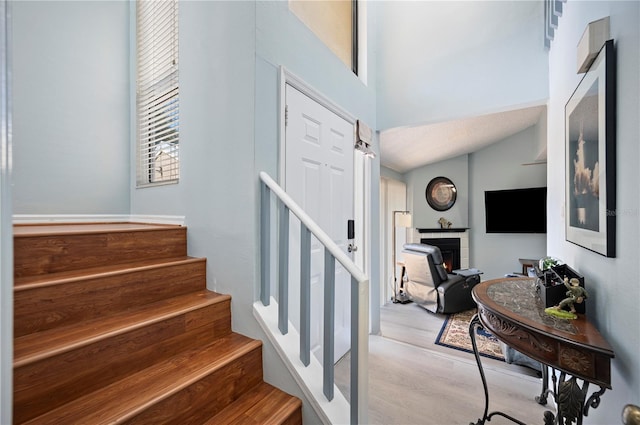 stairway featuring a warm lit fireplace, wood finished floors, and lofted ceiling