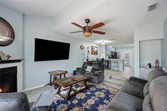 living room featuring visible vents, a tiled fireplace, a ceiling fan, light wood-style floors, and a textured ceiling