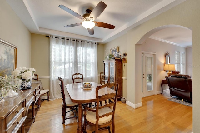 dining area featuring arched walkways, a tray ceiling, light wood-style flooring, a ceiling fan, and baseboards