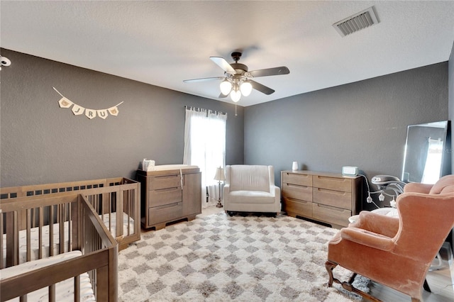 bedroom featuring visible vents, a textured wall, light colored carpet, a ceiling fan, and a nursery area