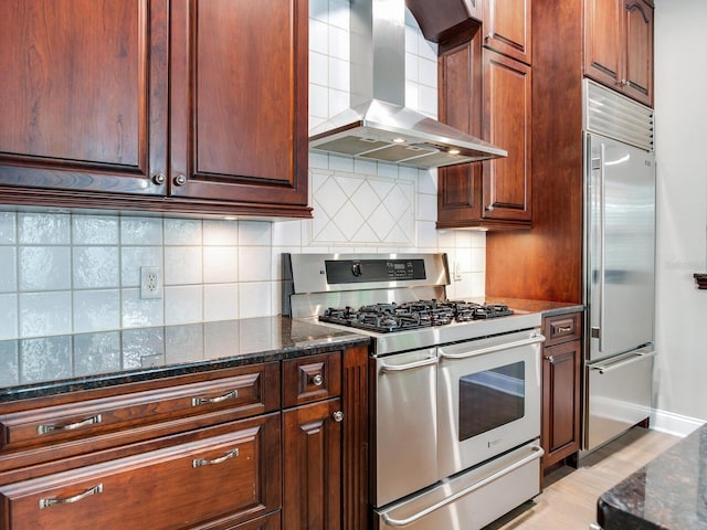 kitchen featuring gas range oven, built in fridge, dark stone countertops, wall chimney exhaust hood, and backsplash