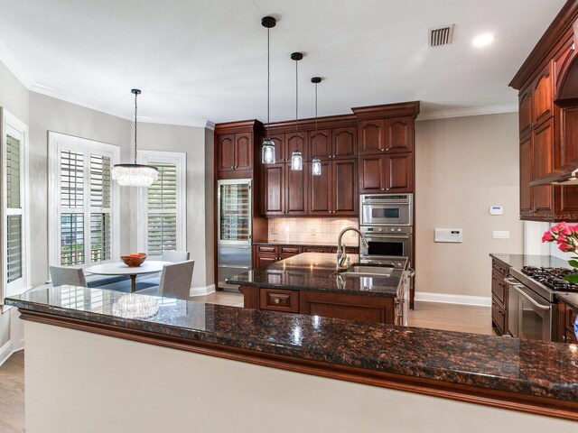 kitchen with decorative backsplash, light hardwood / wood-style flooring, dark stone countertops, an island with sink, and stainless steel appliances