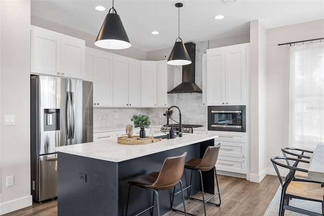 kitchen with a center island with sink, white cabinets, stainless steel appliances, wall chimney range hood, and pendant lighting