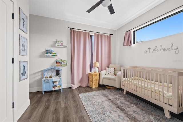 bedroom featuring a crib, a ceiling fan, baseboards, and dark wood-style flooring