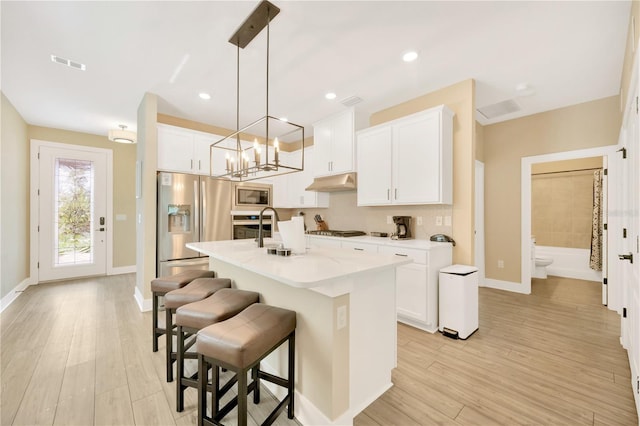 kitchen featuring under cabinet range hood, stainless steel appliances, white cabinets, hanging light fixtures, and a center island with sink