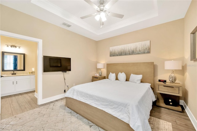 bedroom with light wood finished floors, visible vents, baseboards, ensuite bath, and a tray ceiling