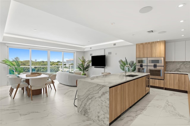 kitchen featuring a raised ceiling, light stone countertops, sink, a center island with sink, and light tile patterned flooring