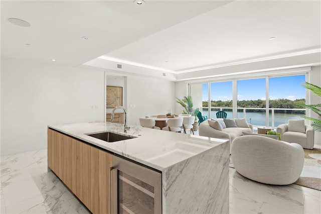 kitchen featuring a kitchen island with sink, light stone counters, wine cooler, sink, and a water view