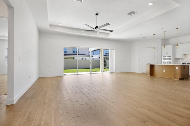 unfurnished living room featuring light wood-type flooring, a tray ceiling, baseboards, and a ceiling fan