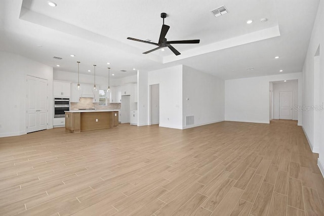 unfurnished living room with a ceiling fan, a tray ceiling, light wood-type flooring, and visible vents