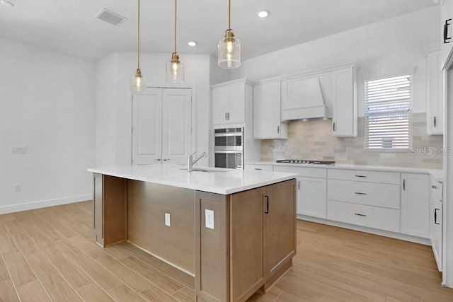 kitchen featuring premium range hood, light hardwood / wood-style floors, white cabinets, and an island with sink