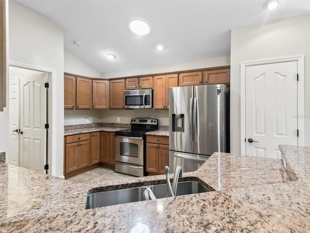 kitchen with lofted ceiling, light stone counters, stainless steel appliances, a sink, and brown cabinetry