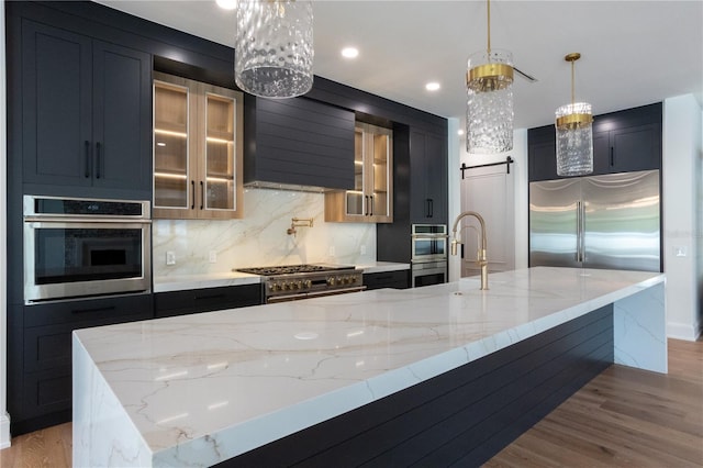 kitchen featuring high end appliances, tasteful backsplash, light wood-style flooring, a barn door, and wall chimney exhaust hood