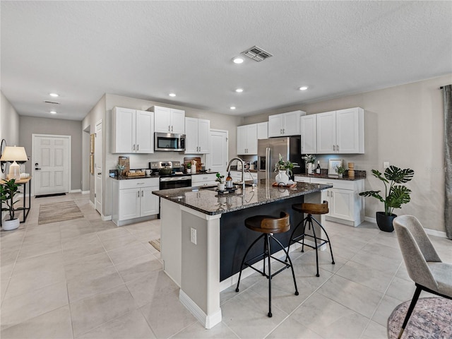 kitchen with stainless steel appliances, white cabinetry, a center island with sink, and dark stone countertops