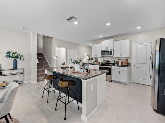 kitchen with a kitchen island with sink, white cabinets, a kitchen bar, stainless steel appliances, and dark stone countertops