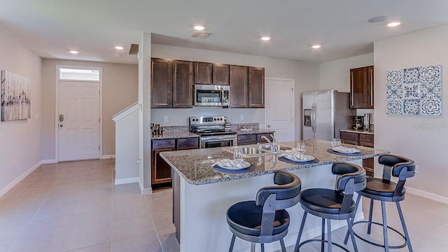 kitchen with stainless steel appliances, an island with sink, stone countertops, light tile patterned floors, and a breakfast bar